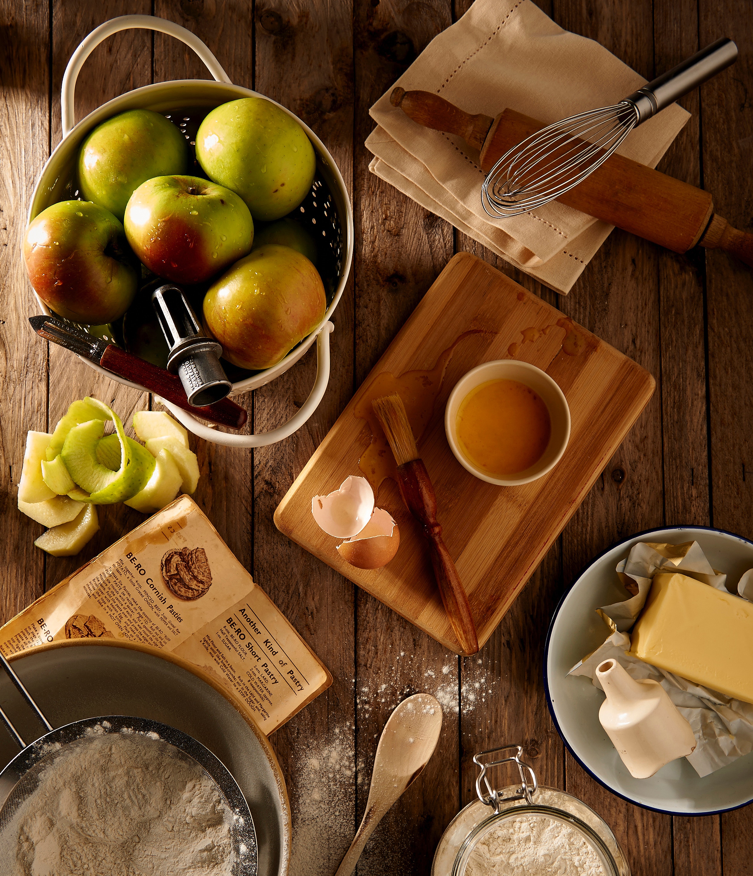 Close up on a kitchen counter showing a recipe book, mixing bowls with white flour in. Apples are in a kitchen strainer, one of them peeled. a wooden chopping board has a small bowl with egg wash in and a pastry brush lays next to it. A bowl of butter appears towards the right of the page. This images shows that someone is perhaps making an apple pie.